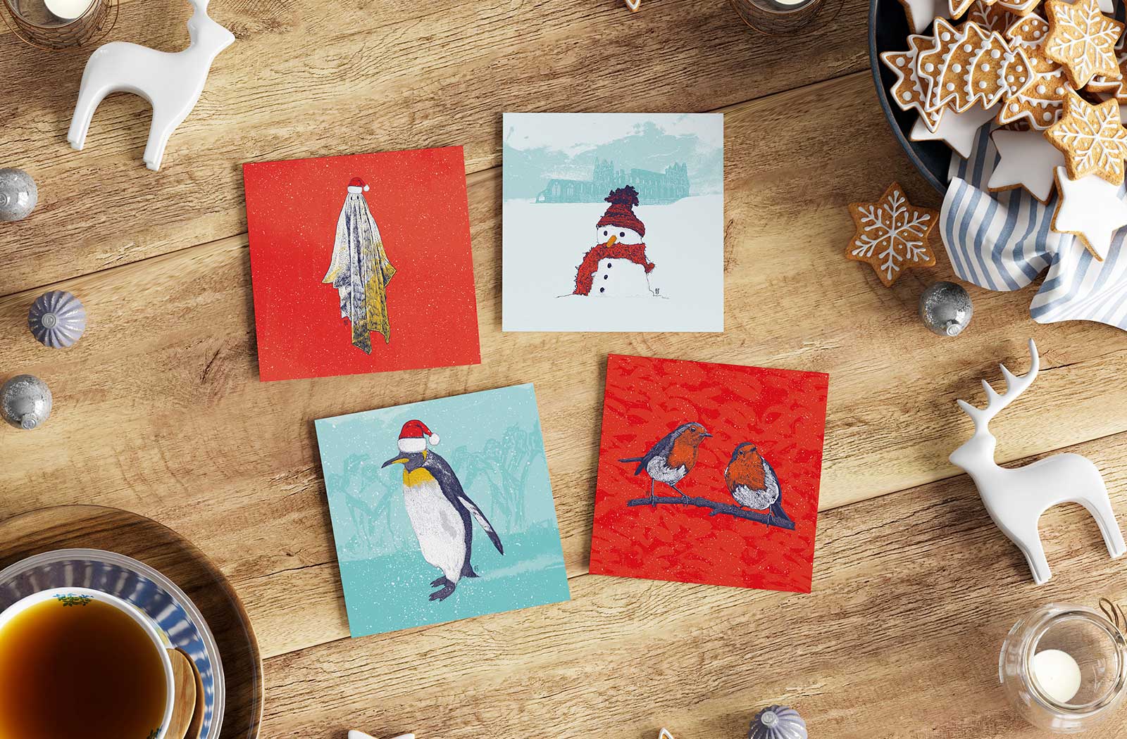 Christmas cards with a ghost, snowman, penguin, and robins in Santa hats, surrounded by holiday decor and cookies.